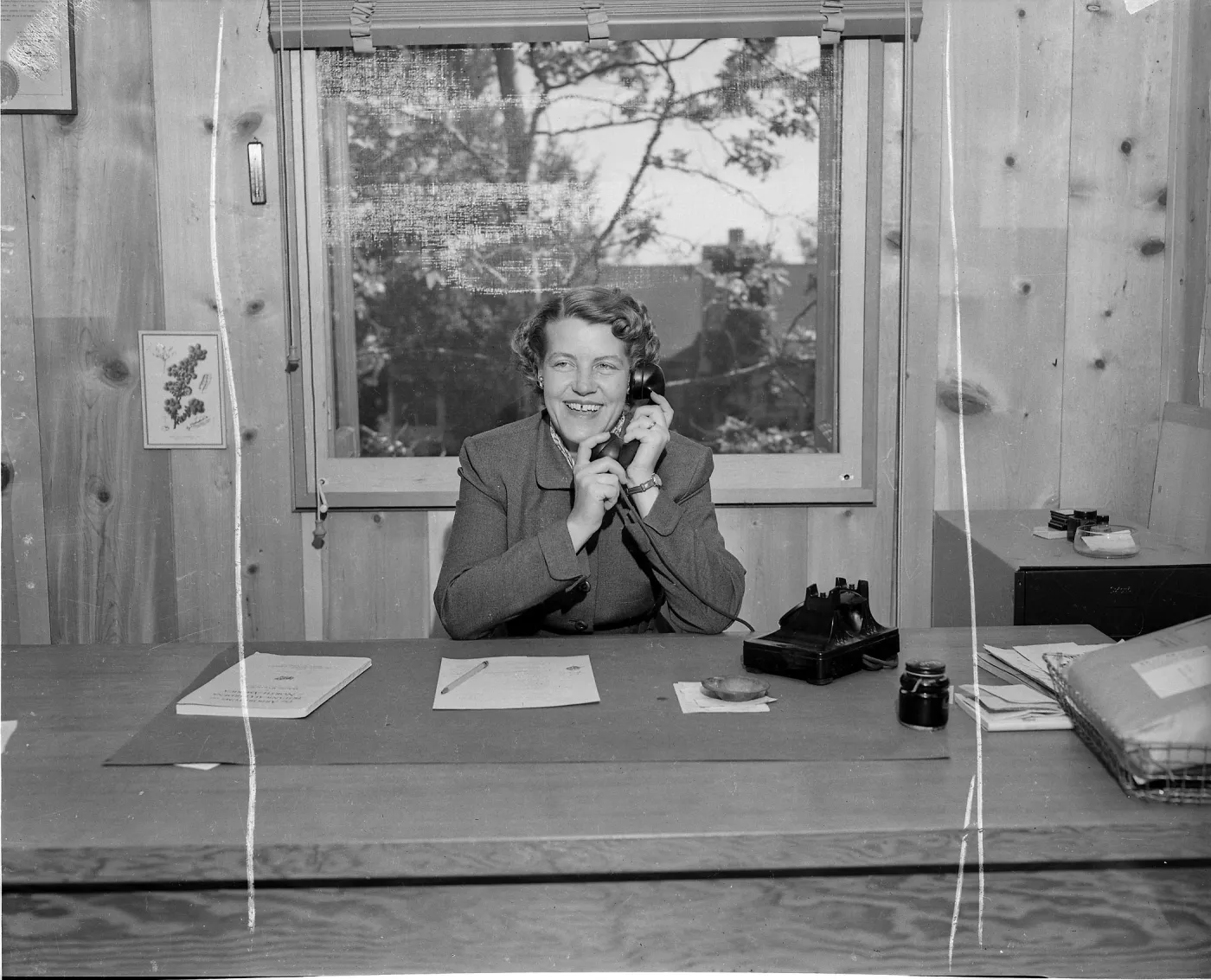 Black-and-white photo of a smiling woman in a professional jacket seated at a desk, holding a telephone receiver in a wood-paneled office with papers and a rotary phone on the desk.
