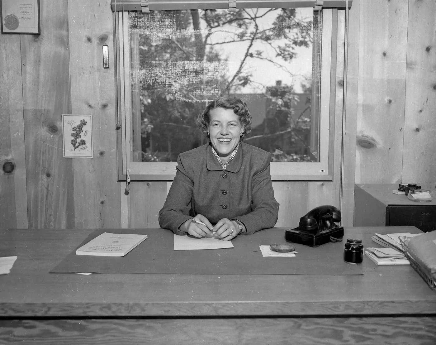 A black and white photo of a smiling woman at a desk in a wood paneled office with a rotary phone next to her.