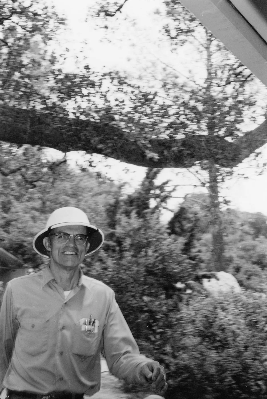 Black-and-white photo of a smiling man in glasses and a brimmed hat, wearing a button-up shirt, standing outdoors with trees and shrubs behind him.