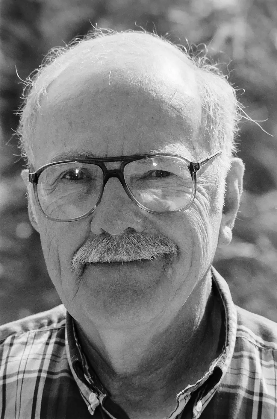 Black and white close-up of an older man wearing glasses and a plaid shirt, looking at the camera.