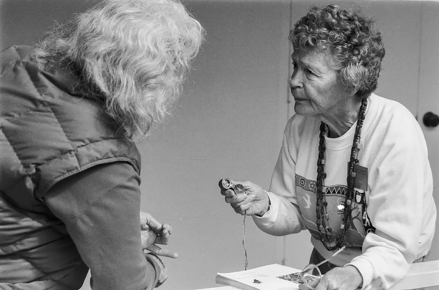 a black and white photo of an older woman with white hair and their back facing the camera talking to an older woman sharing an activity.