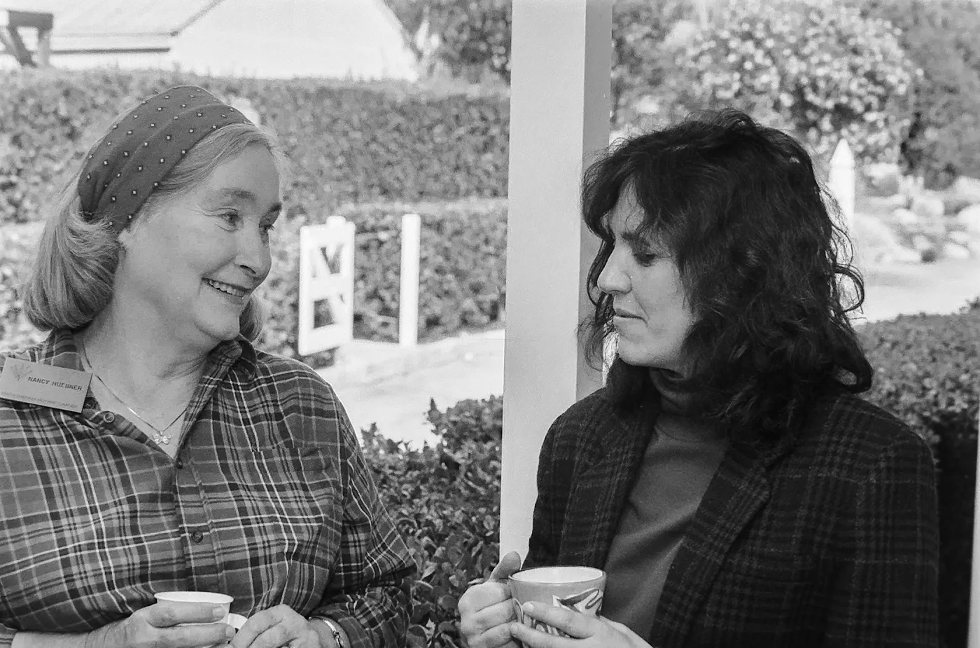 Older woman in a headscarf, nametag and plaid shirt talking to a younger woman in a jacket outdoors while they both hold cups.