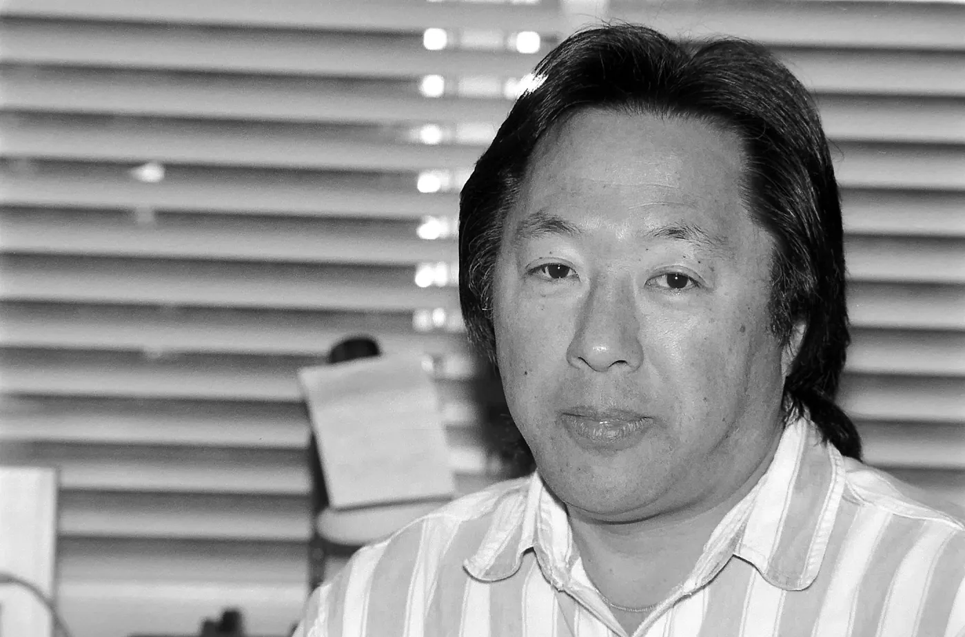 a black and white photo of a middle aged Asian man in a striped shirt with office blinds behind him.