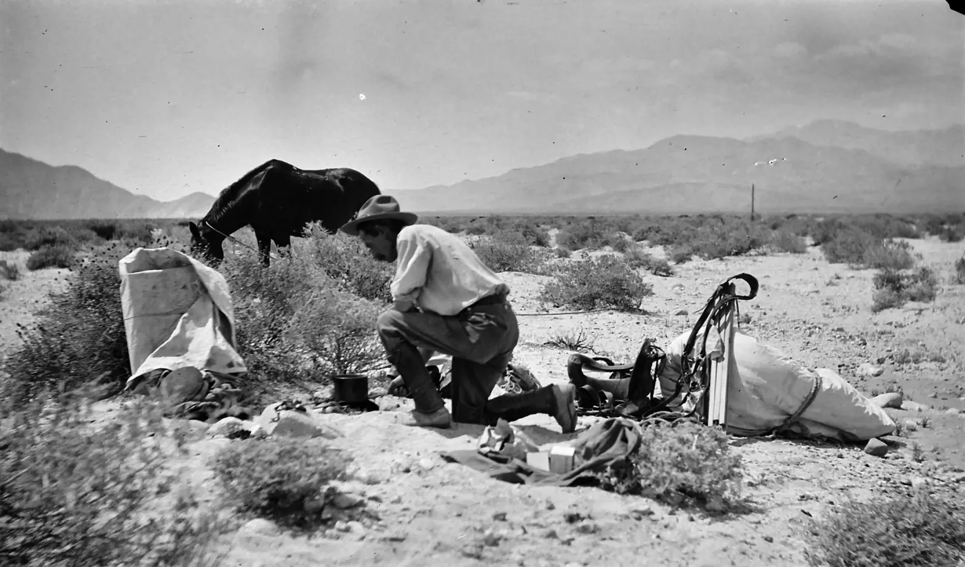Black and white photograph of a man in a hat kneeling in a desert landscape next to a pile of equipment with a black horse grazing behind him.