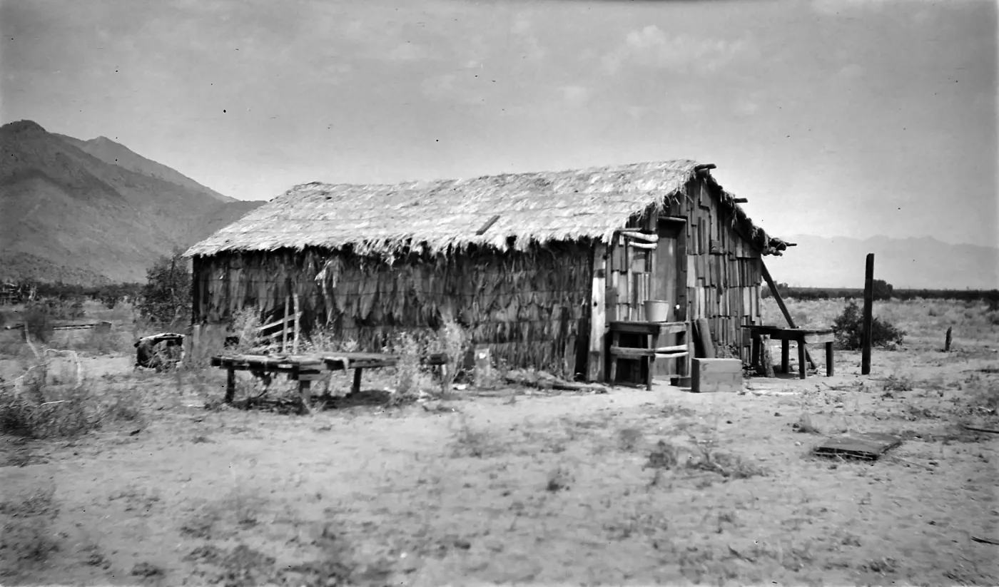 Black and white photograph of a wooden structure with a thatched roof in a desert setting with mountains in the distance.