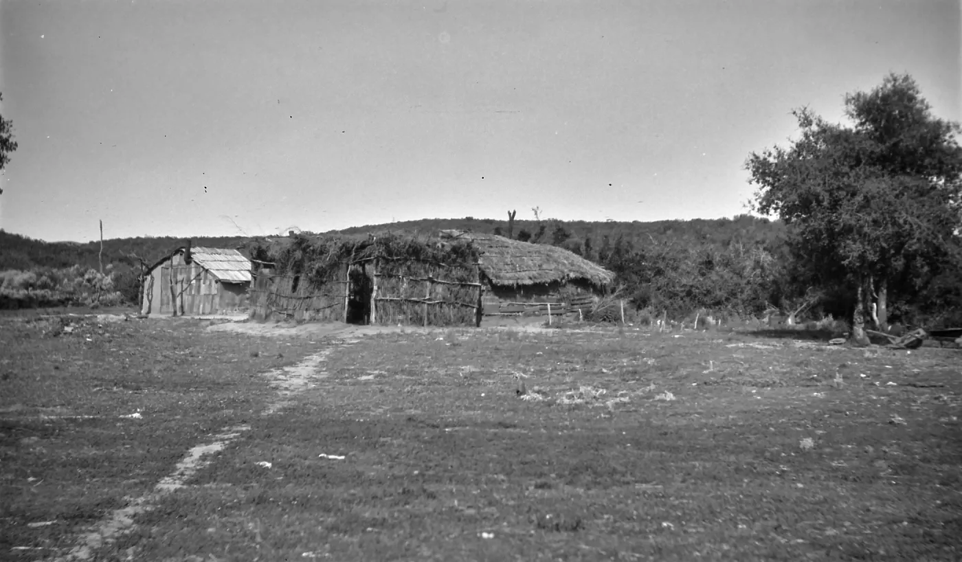 Black and white photograph of a low thatched structure with an open field in front of it and a path worn through the grass.
