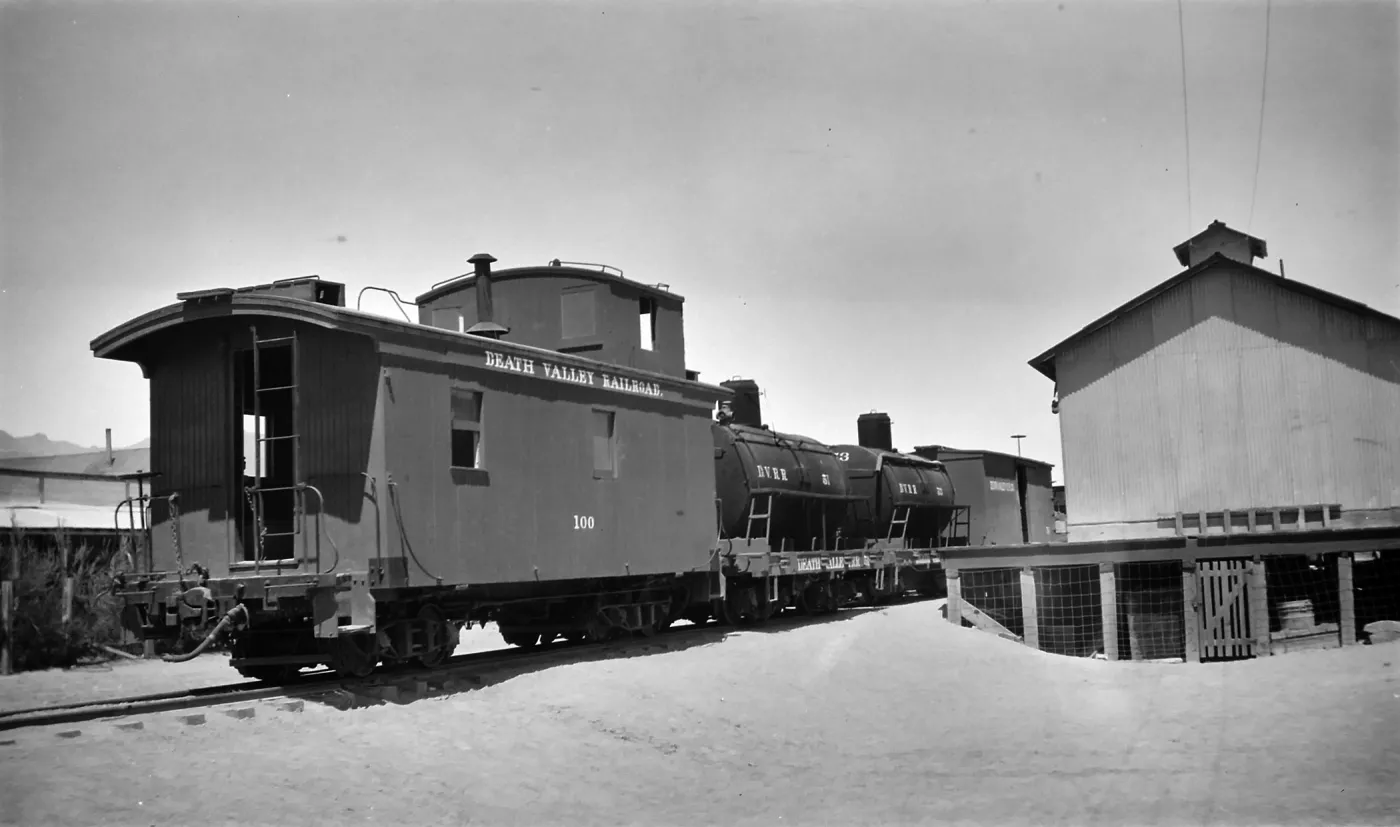Black and white photograph of a train station, with a wooden building and three black train cars visible. The first car says Death Valley Railroad.