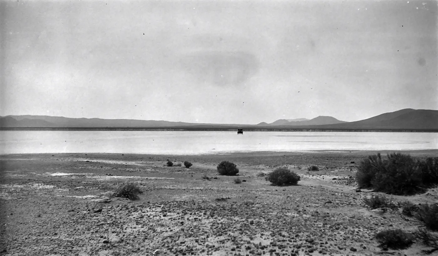 Black and white photograph of a black car on a wide white salt flat with hills in the distance.
