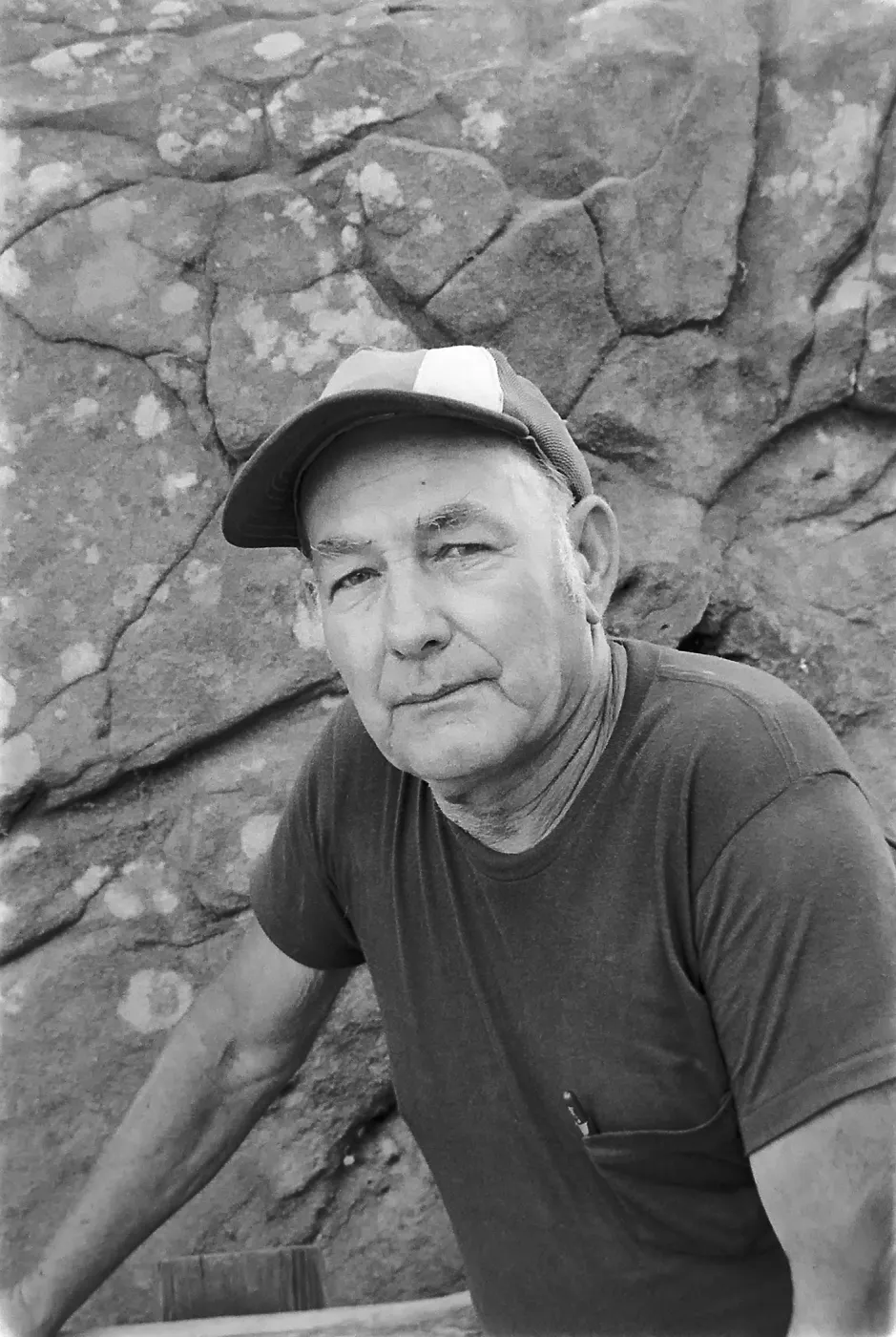Black and white photograph of a man in a short sleeved shirt and baseball cap looking at the camera against a rock wall background.
