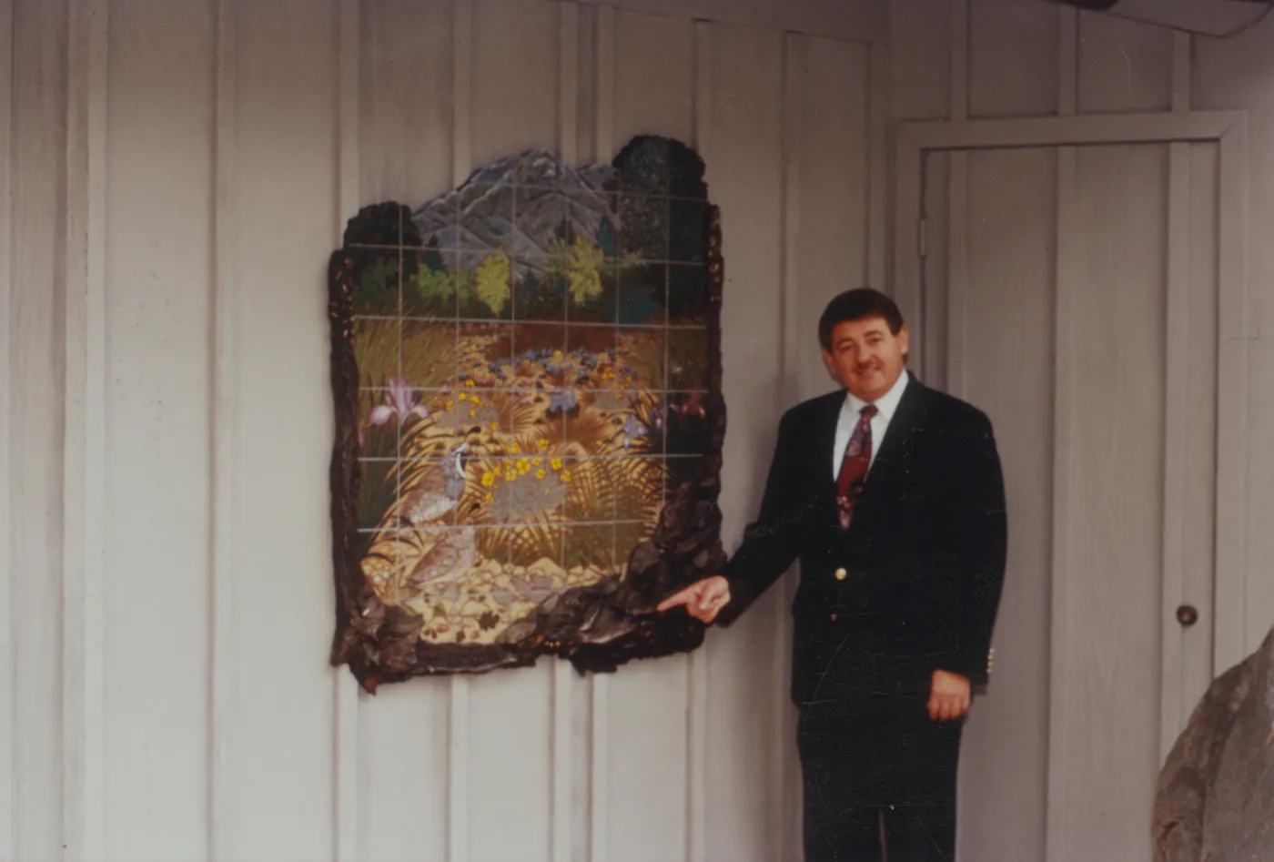 A man in a suit and red tie points at an irregularly shaped mosaic hanging on a white wood paneled wall. The mosaic depicts two California quails in a meadow of long, yellow and dull green grasses and yellow, pink, and pale blue flowers, in front of a background of trees and mountains. A grid pattern is overlaid the mosaic image, and the edges of the piece are deep brown wood carved in the shapes of leaves. 