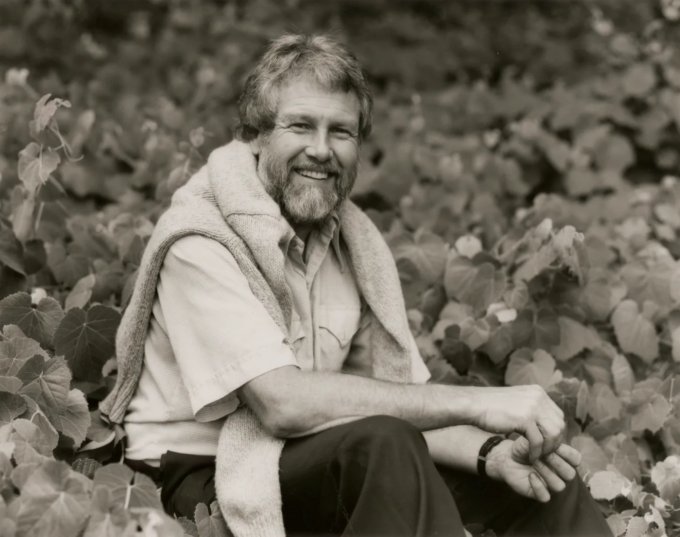 A black and white photo of a bearded man wearing a light-colored shirt and dark slacks with a light-colored sweater over his shoulders. He sits in a field of vines with many heart-shaped leaves. 