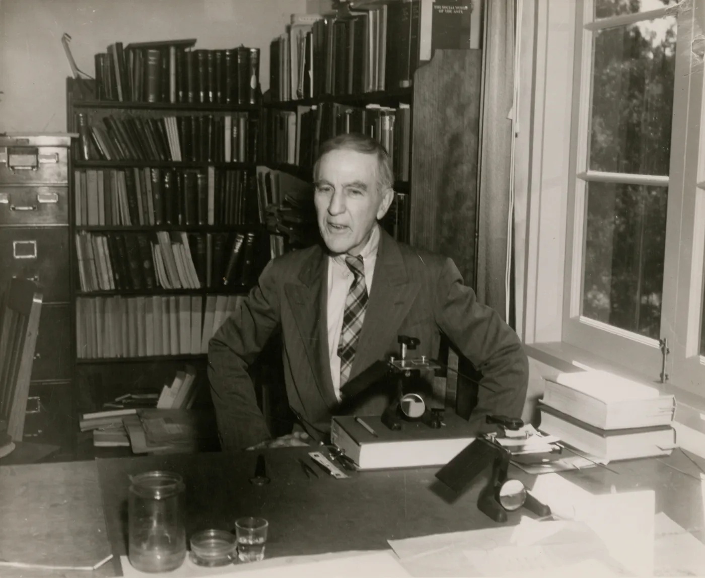 A black and white photo of a man in a suit and plaid tie sits at a desk in front of shelves full of books. 