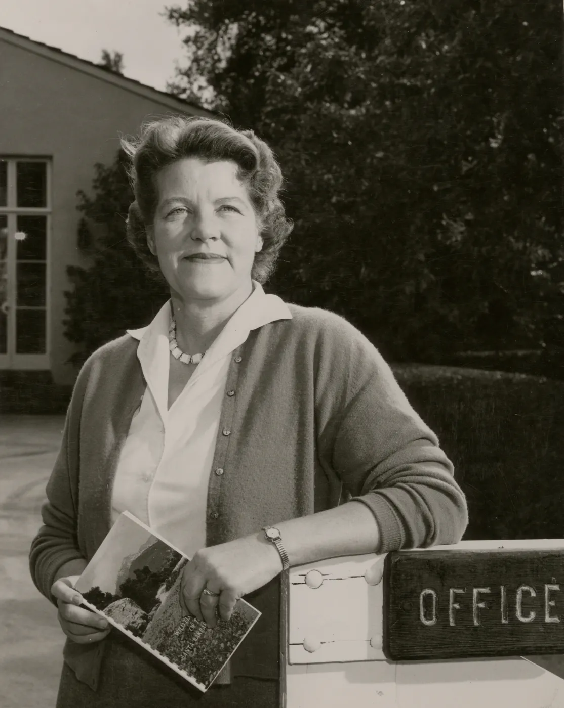 A black and white photo of a woman in a blouse posing in front of a building with neatly trimmed hedges. She holds a booklet depicting a field in front of a forest and mountain and one of her arms rests upon a wooden fence gate with a sign reading "Office" cutoff at the edge of the image.