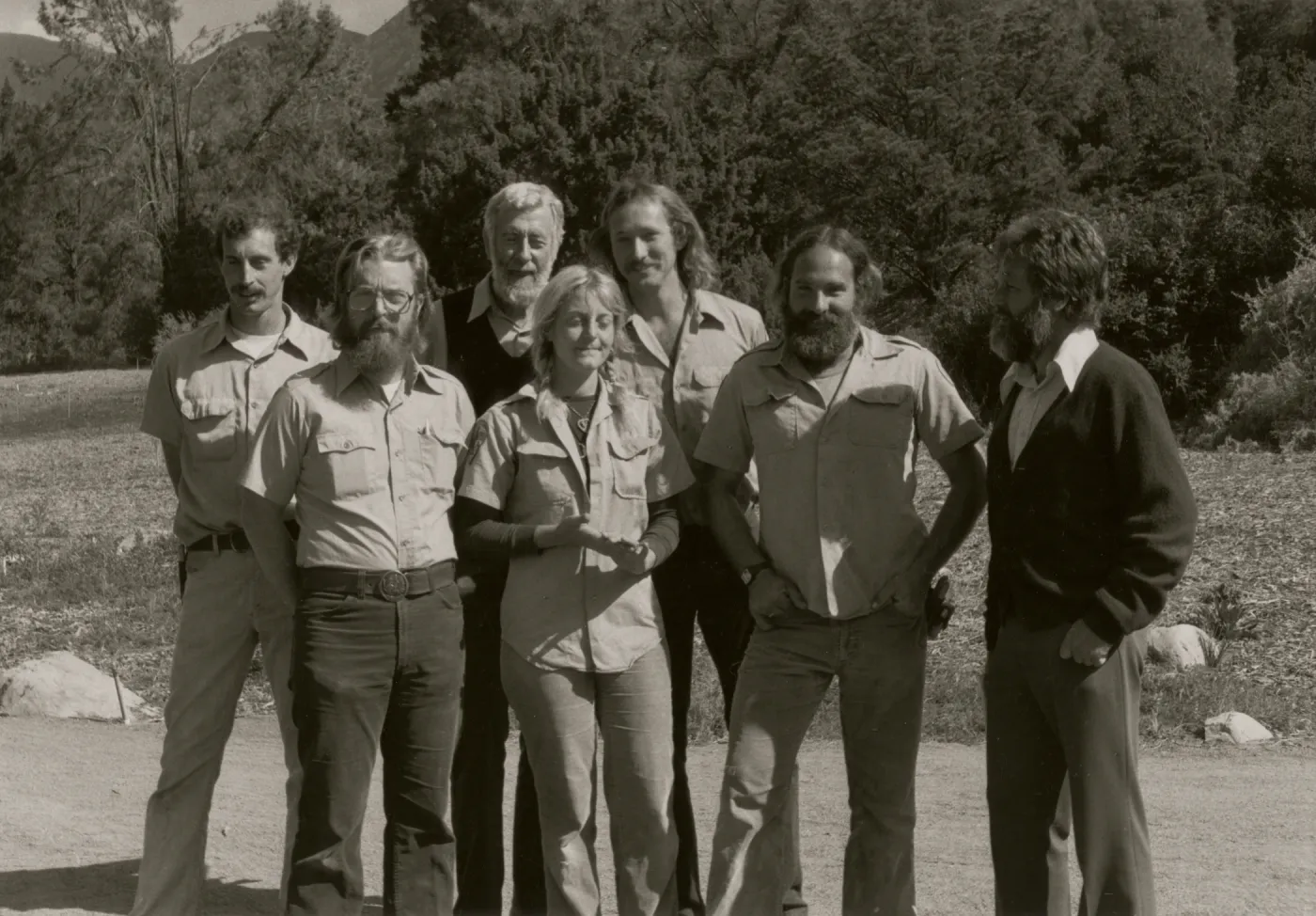 A black and white photo of seven people wearing slacks and button-up shirts stand in a rocky field at the edge of a copse of trees.