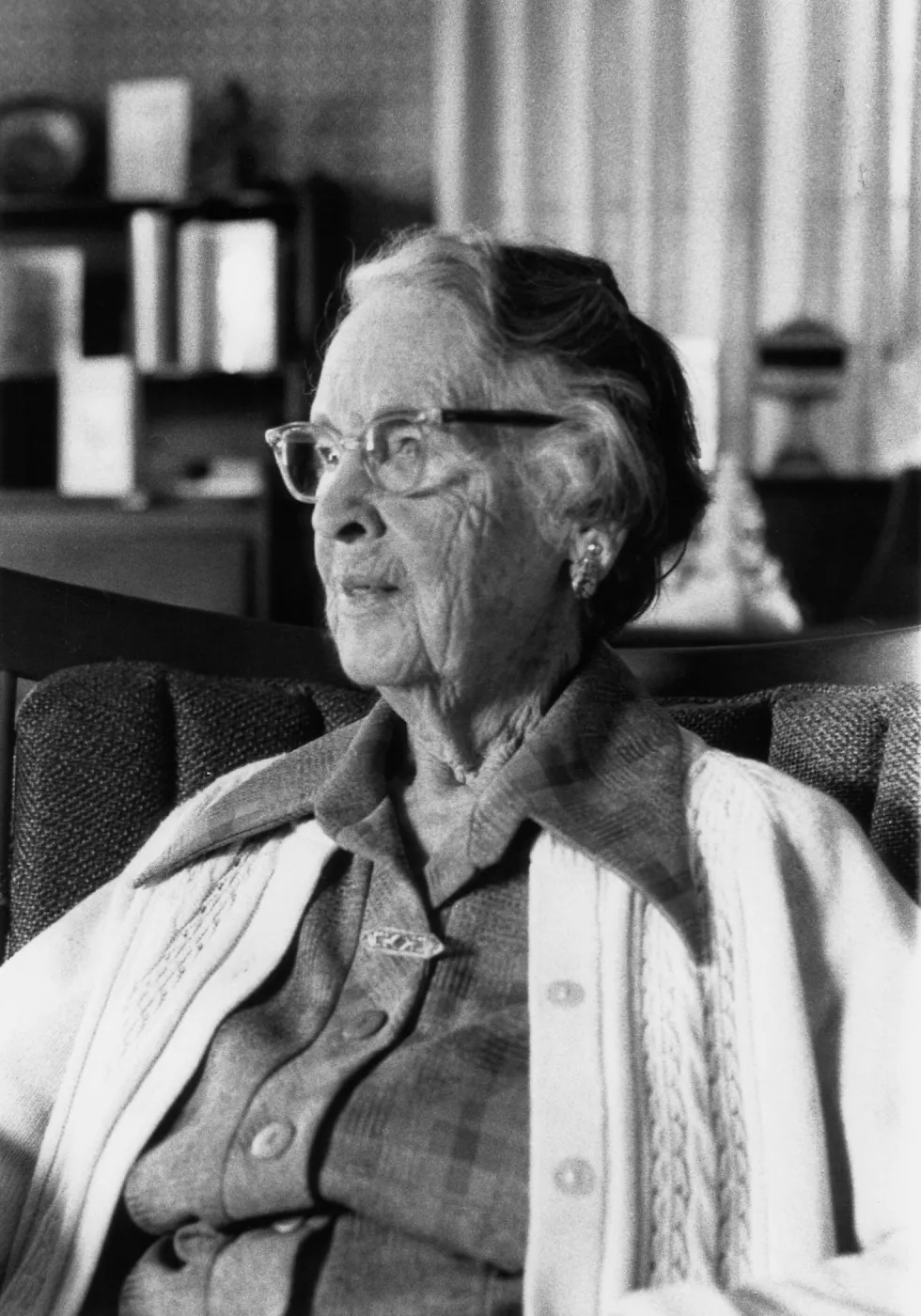 Black and white photograph of woman sitting in chair looking out in distance wearing button up cardigan and glasses.