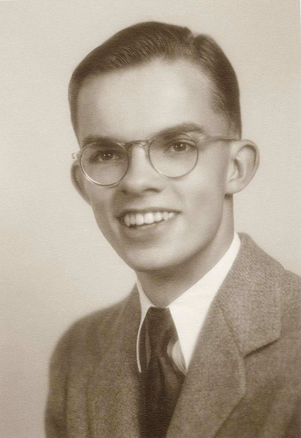 Black and white photograph of young male posing in suit and tie with glasses. 