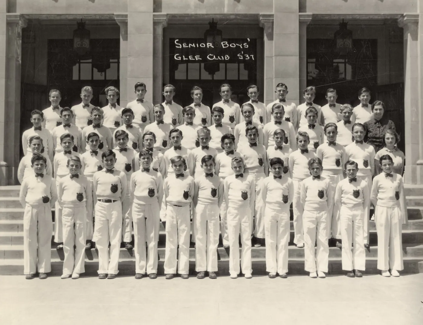 Black and white photograph shows four rows of high school boys in white uniforms standing in front of building with poster. 