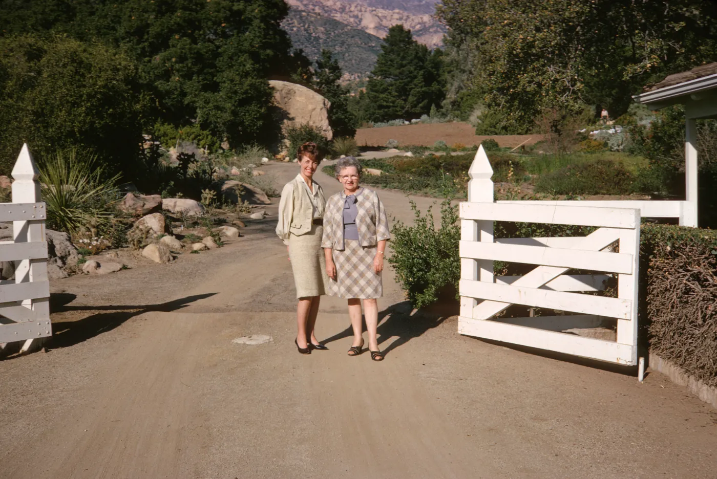 Two women, one older and one younger, in pale colored suits stand, smiling on a gravel road in an open white fence gate. The road behind them is lined with rocks and trees, with mountains and a large boulder visible in the background.