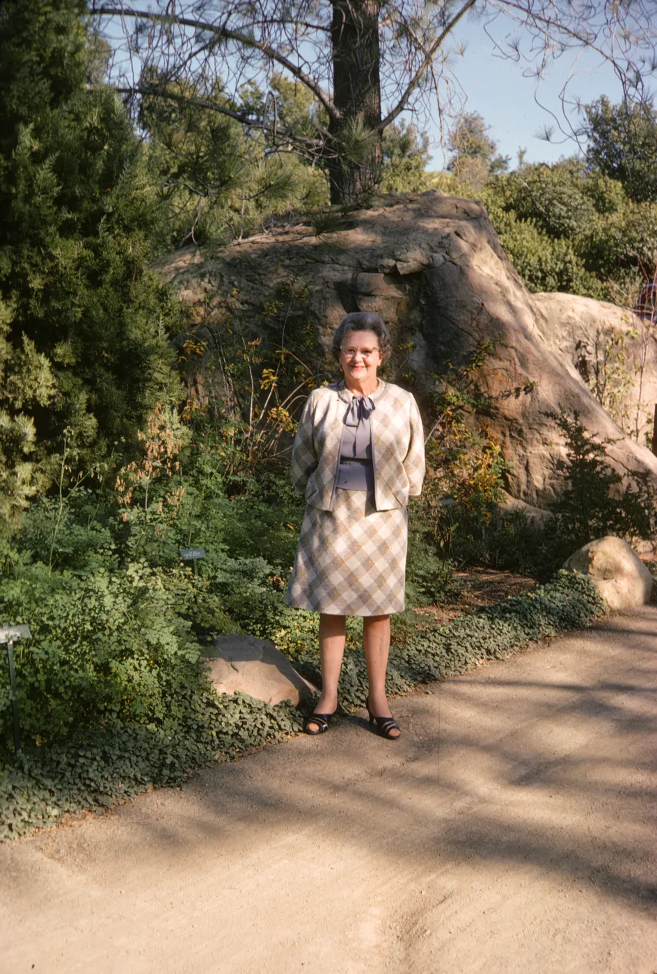An older woman in a pale suit smiles at the edge of a paved road. Next to her at the edge of the road is a tall evergreen tree surrounded by low greenery and behind her is a large boulder.