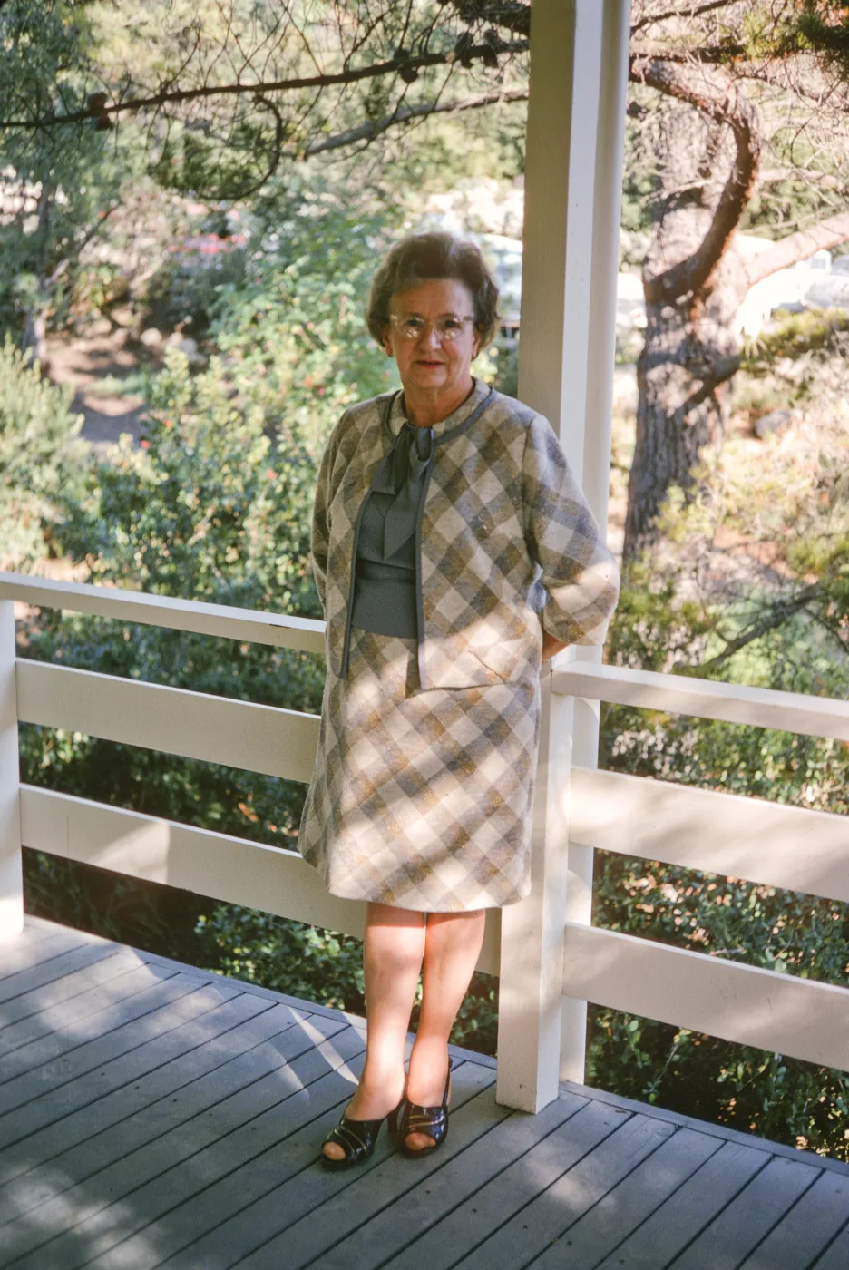 An older woman in a pale, checkered suit stands on a shady porch against a white, wooden railing. 