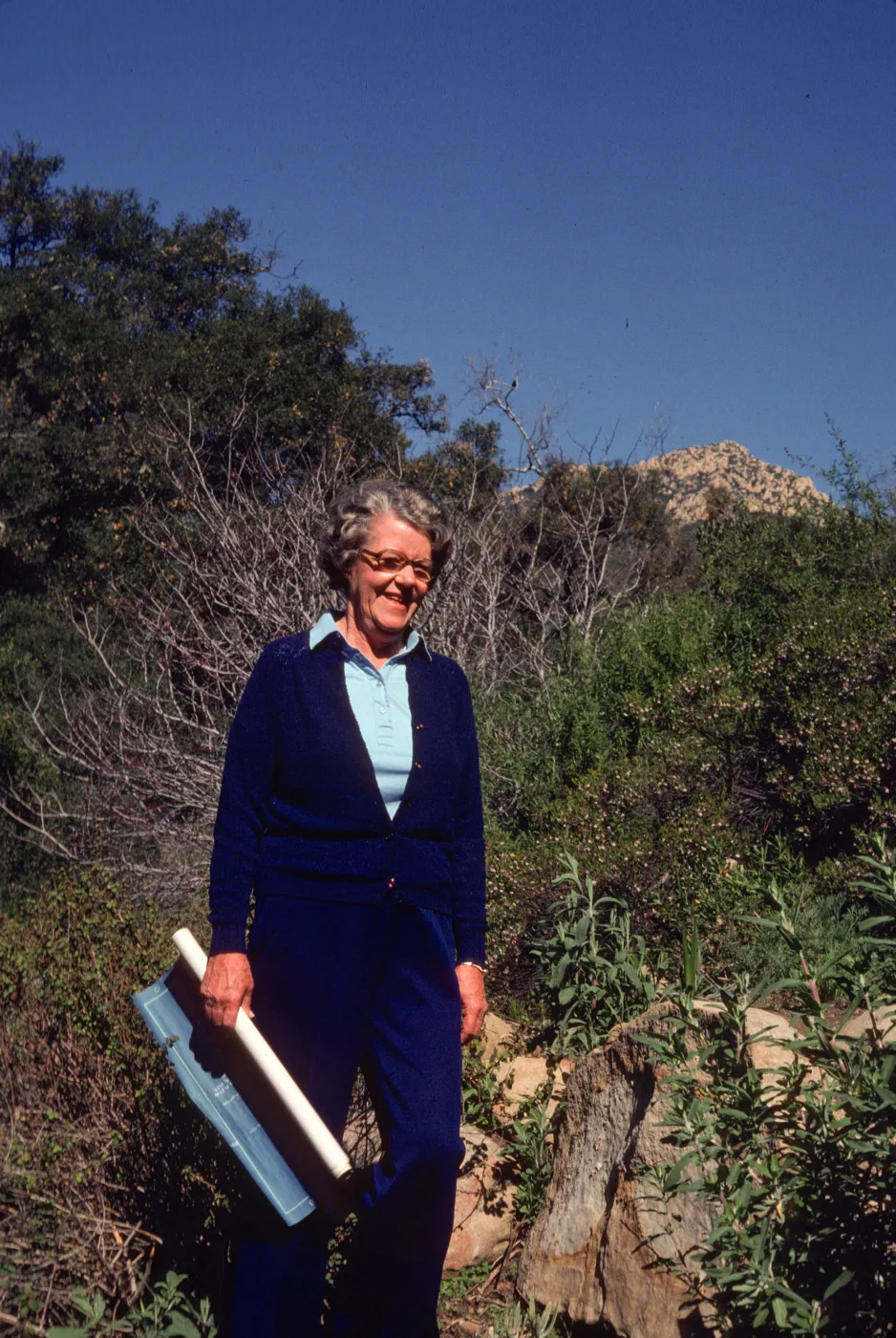 An older woman dressed in royal blue smiles in front of a rocky hill coated in short vegetation and bare-branched bushes. A rolled blueprint is held in her right hand. 
