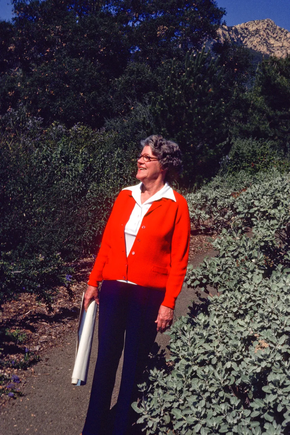 An older woman in a bright orange-red sweater stands on a path flanked on both sides by tall bushes, smiling into the distance. She holds a rolled set of blueprints in her right hand. 