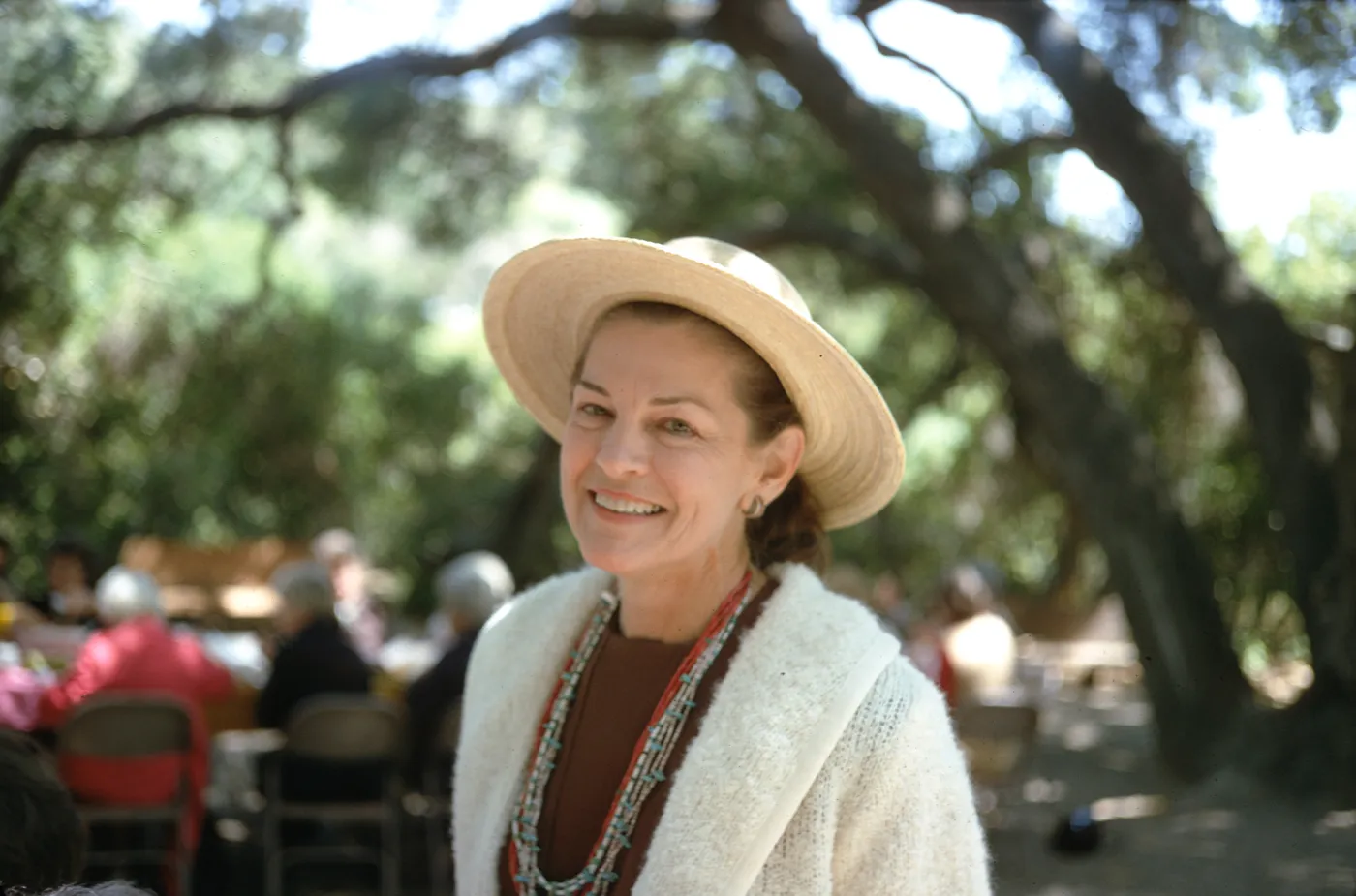 A woman in a straw hat, white cardigan, and numerous beaded necklaces smiles at the camera. In the background, several people sit at an outdoor table beneath a large double-trucked tree.