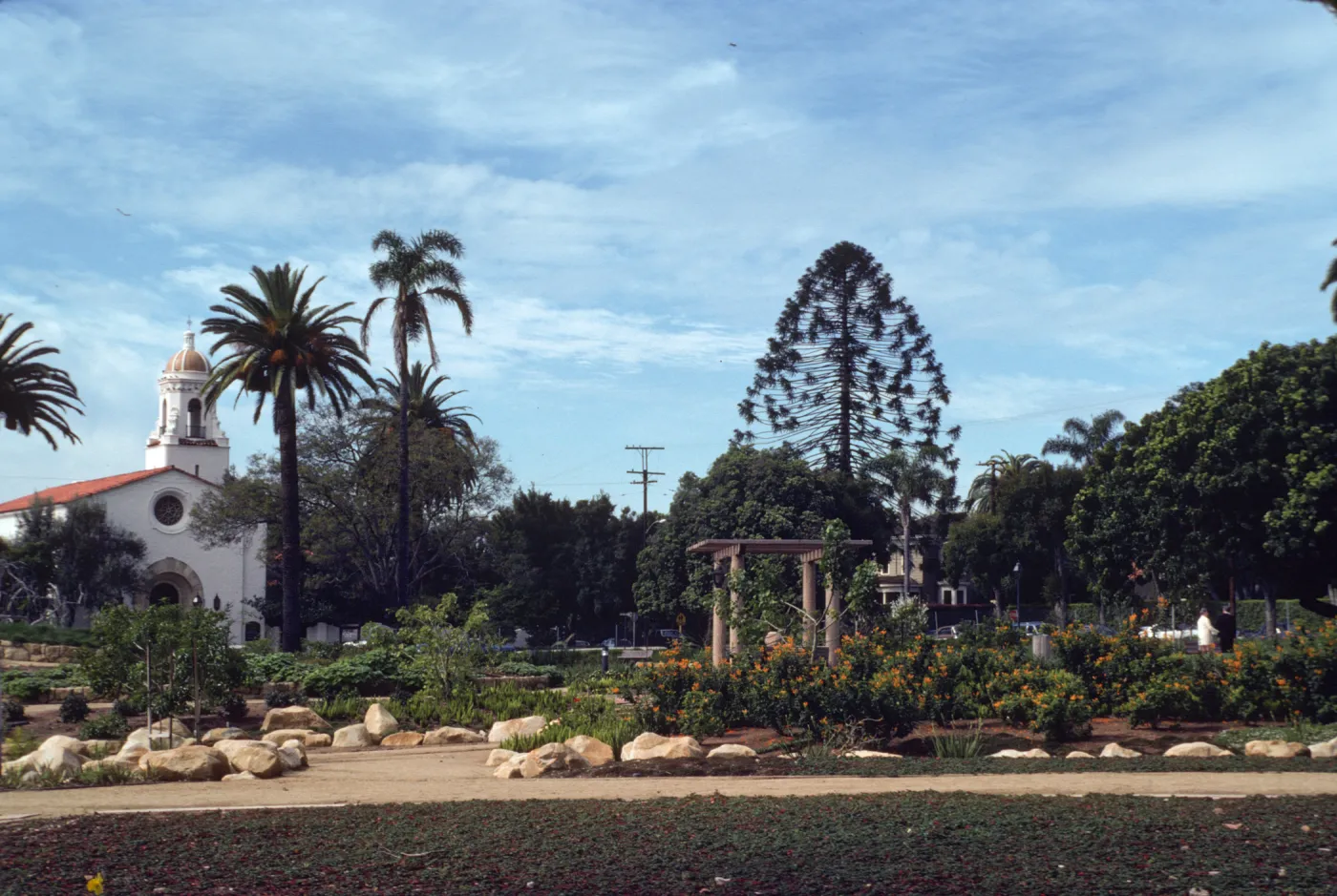 A variety of plants grow in beds lines with large stones and divided by footpaths. A large, white building with a golden-domed steeple is visible at the edge of the garden.
