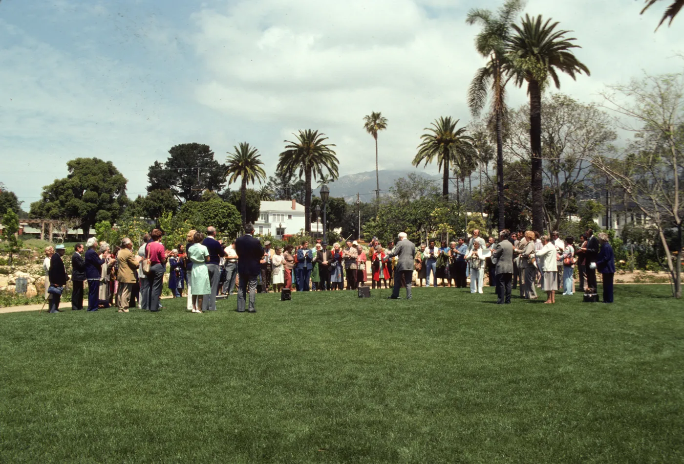 A large group of well-dressed people stand in a semicircle in a grassy yard. An older man in a grey suit stands in the center of the semi-circle.