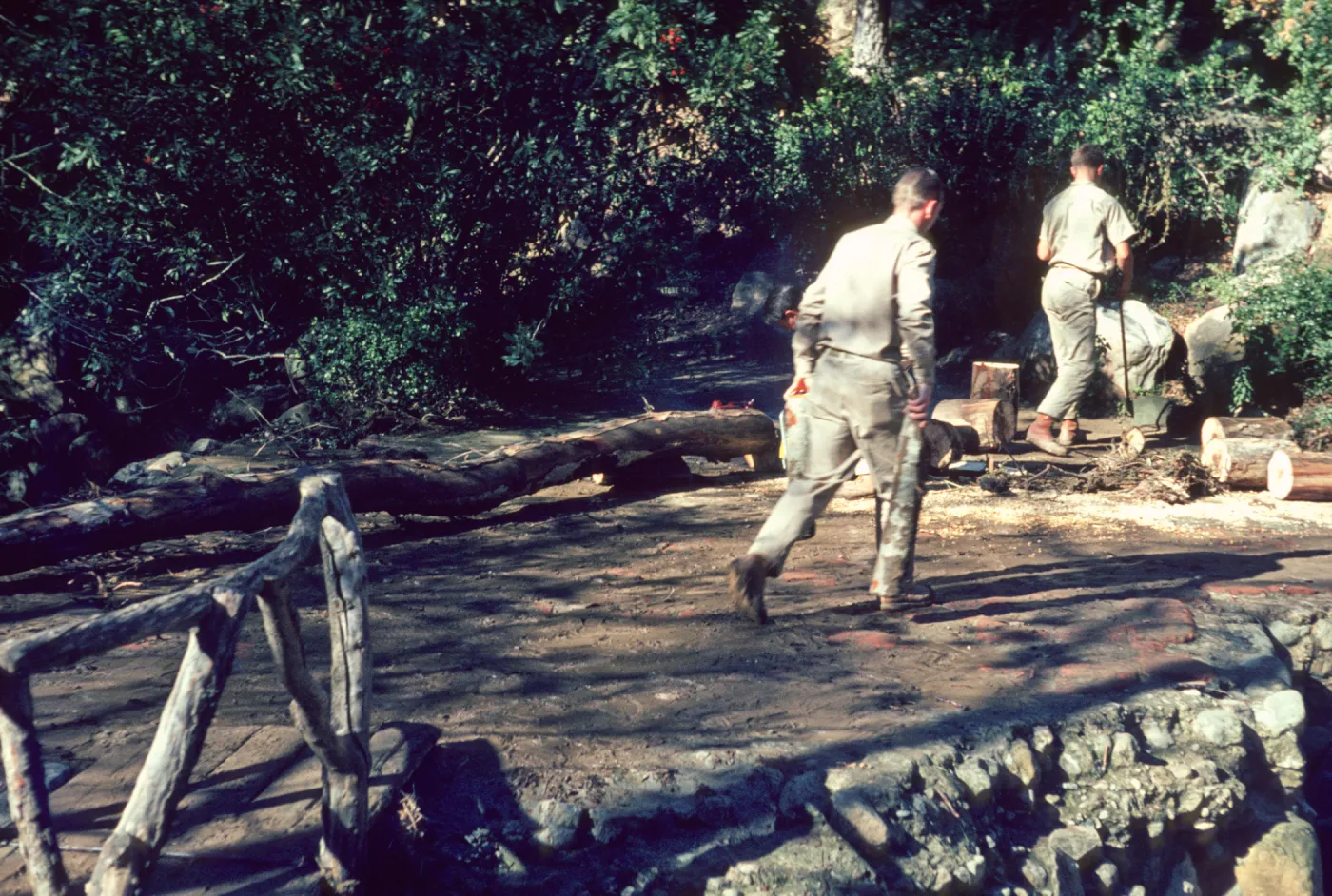Two men in khaki work clothes chopping wood on muddy, stony ground at the edge of a wooded area. A rustic wooden bridge leads into the area.