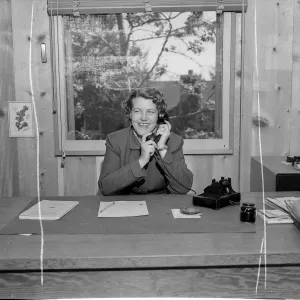 Black-and-white photo of a smiling woman in a professional jacket seated at a desk, holding a telephone receiver in a wood-paneled office with papers and a rotary phone on the desk.