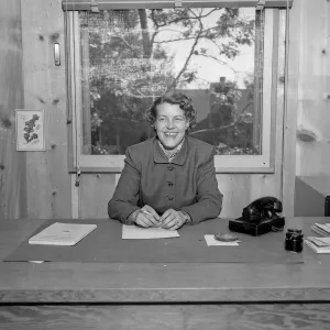 A black and white photo of a smiling woman at a desk in a wood paneled office with a rotary phone next to her.