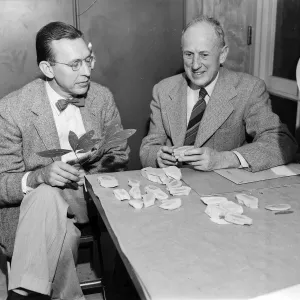 Black and white photo of two men in suits seated at a table examining fossils while the man on the left holds a plant cutting.