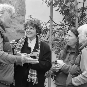 Four women standing outdoors in conversation, two holding cups while another gestures with her hands.
