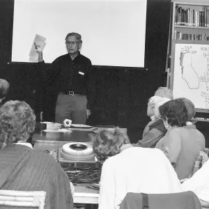 A black and white photo of a man holding up a piece of paper in front of a group of people sitting at a table with a whiteboard to the right.