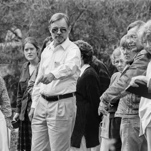 A black and white photo of a man in sunglasses pointing and speaking to a group of people outdoors who stand in a line listening.