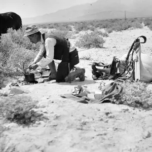 a black and white photo of a man kneeling on desert ground stirring a pot over a small fire with packs and gear spread nearby and a horse grazing in the background.