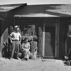 A black and white photo of a man in a cowboy hat standing and two women sitting outside an old wooden building. 