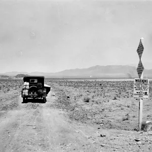 Black and white photograph of a black car driving down a dirt road into the desert past a sign that says Warning.