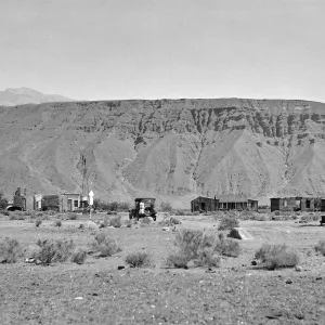 Black and white photograph of an abandoned town of wooden buildings with a mesa in the background.