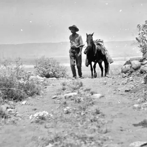 Black and white photograph of a man in a hat walking a black horse down a rock-strewn desert pathpath.