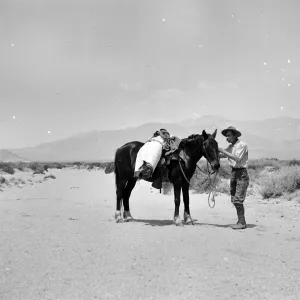 Black and white photograph of a man in a hat standing next to a black horse with a white blanket rolled on its back with hills in the distance.