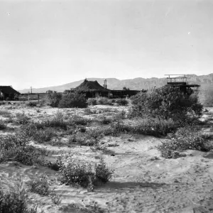 Black and white photogaph of a group of one-story buildings in a desert setting with brush and hills in the background.