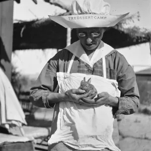 Black and white photograph of a woman in sunglasses and a large white hat reading The Desert Camp holding a small rabbit in her hands.