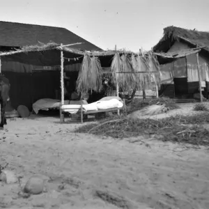 Black and white photograph of a small wooden structure with an open-air room containing two cots with white blankets. There are palm fronds hanging from the roof, and a person is dismounting a black horse in front of the building.