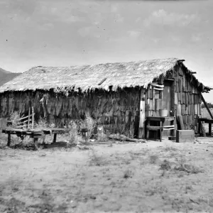 Black and white photograph of a wooden structure with a thatched roof in a desert setting with mountains in the distance.