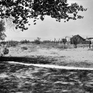 Black and white photograph of an irrigation channel in the foreground and a vineyard, wooden structure, and hills in the background.