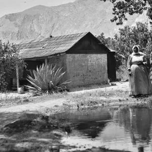 Black and white photograph of a man in a hat and a woman in a dress and head covering next to a pool of water, with a wooden house, trees, desert plants, and mountains in the background.