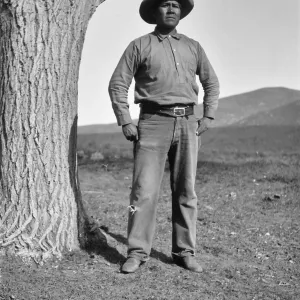 Black and white photograph of a man in a hat, buttoned shirt, and jeans next to a tree with a serious expression.