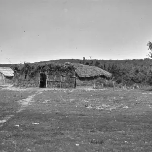 Black and white photograph of a low thatched structure with an open field in front of it and a path worn through the grass.